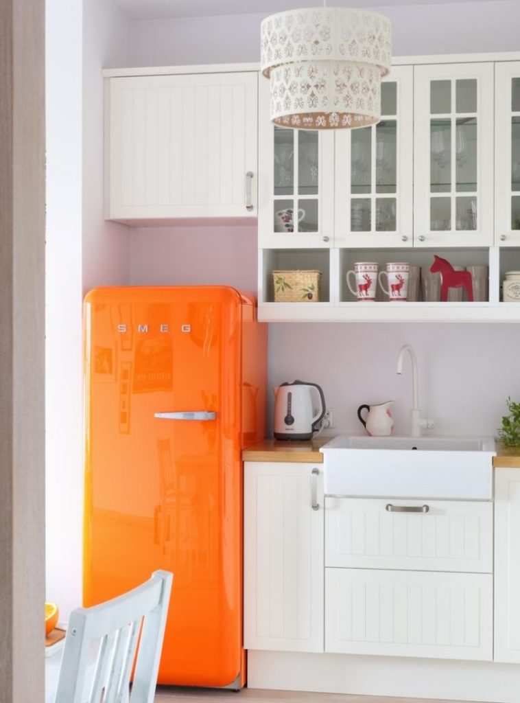 Oranžs ledusskapis virtuvē ar baltām mēbelēm
